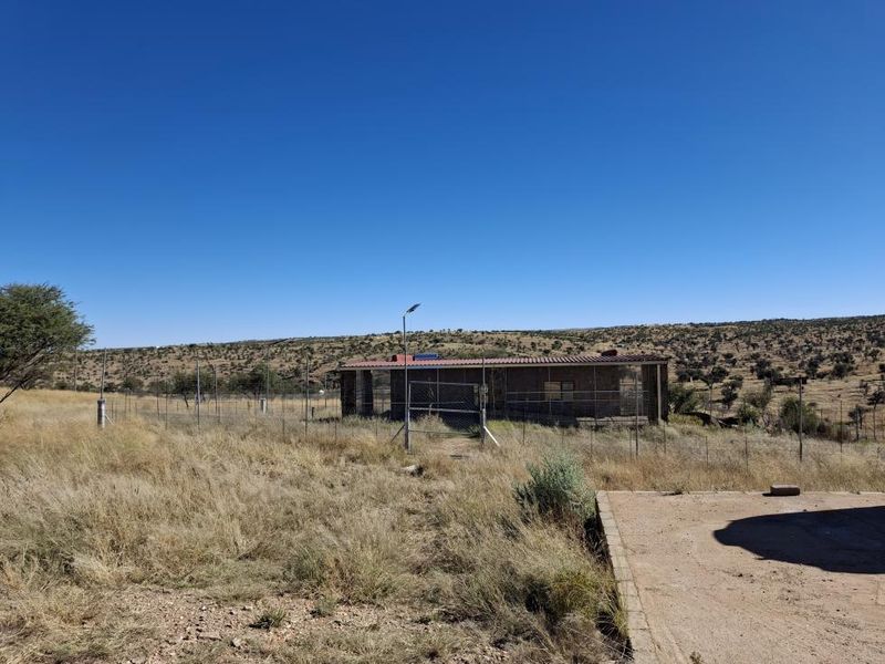 Rear view of a small holding property featuring a fenced perimeter, metal roof structure, and surrounding dry landscape.