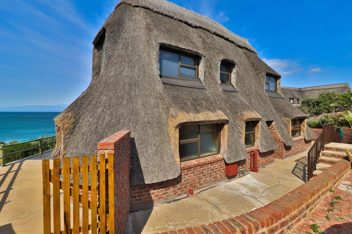A coastal property featuring a distinctive thatched roof, exposed brick facade, and ocean views, set within a landscaped garden area.