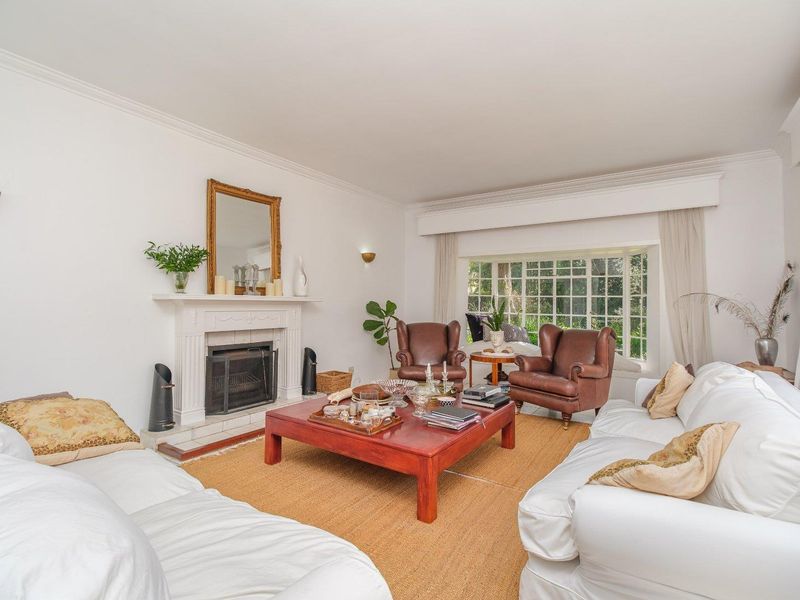 A bright and inviting living room featuring a classic fireplace with a mantelpiece, crown molding, and a large bay window overlooking the garden.