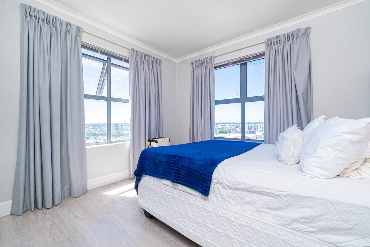 A bright bedroom featuring light wood-look flooring, neutral walls, and large windows offering expansive city views.
