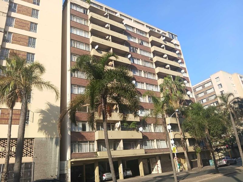 A multi-story apartment building featuring a brick facade, multiple balconies, and ground-level parking facilities in an urban setting.