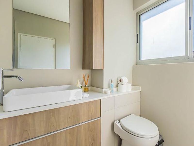 Modern bathroom featuring a vessel sink, wood-finish vanity cabinet, wall-mounted mirror, and a clean, neutral aesthetic with natural light from a sliding window.