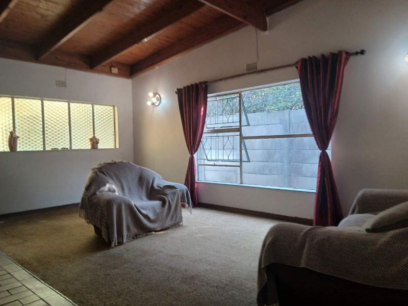 Spacious living room featuring exposed timber ceiling beams, large windows with security bars, and wall-mounted lighting, offering a blank canvas for renovation.