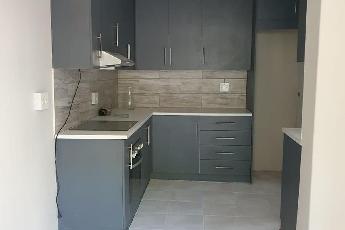A compact, modern kitchen featuring dark grey cabinetry, light-toned tiled splashbacks, a built-in oven and hob, and neutral floor tiles.