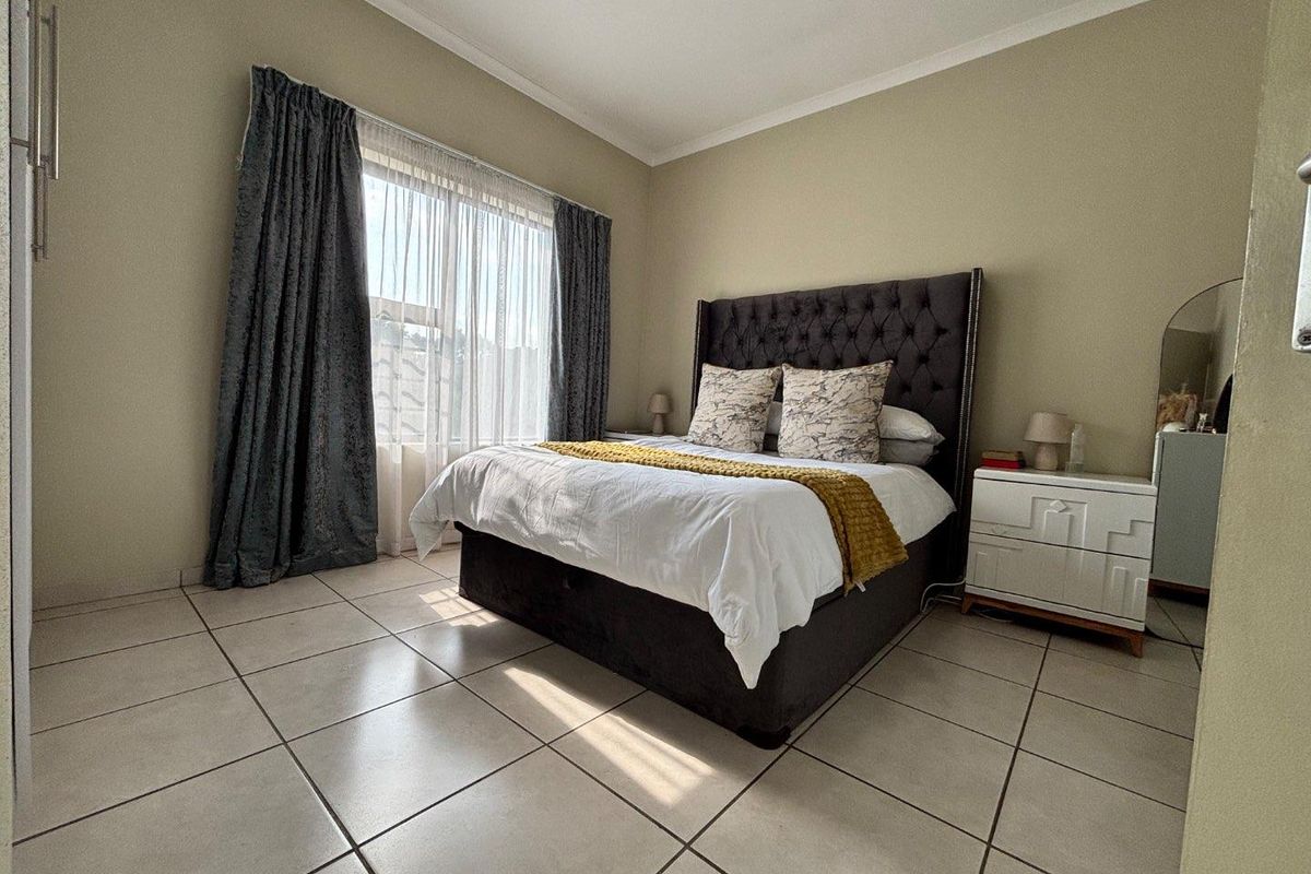 A well-lit bedroom featuring neutral wall tones, large format floor tiles, and a window with curtain tracks.