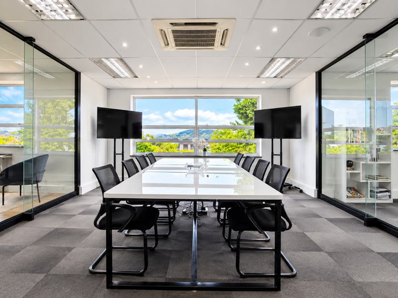 A modern, professional boardroom featuring glass partition walls, a suspended ceiling with integrated air conditioning, and carpeted flooring with ample natural light.
