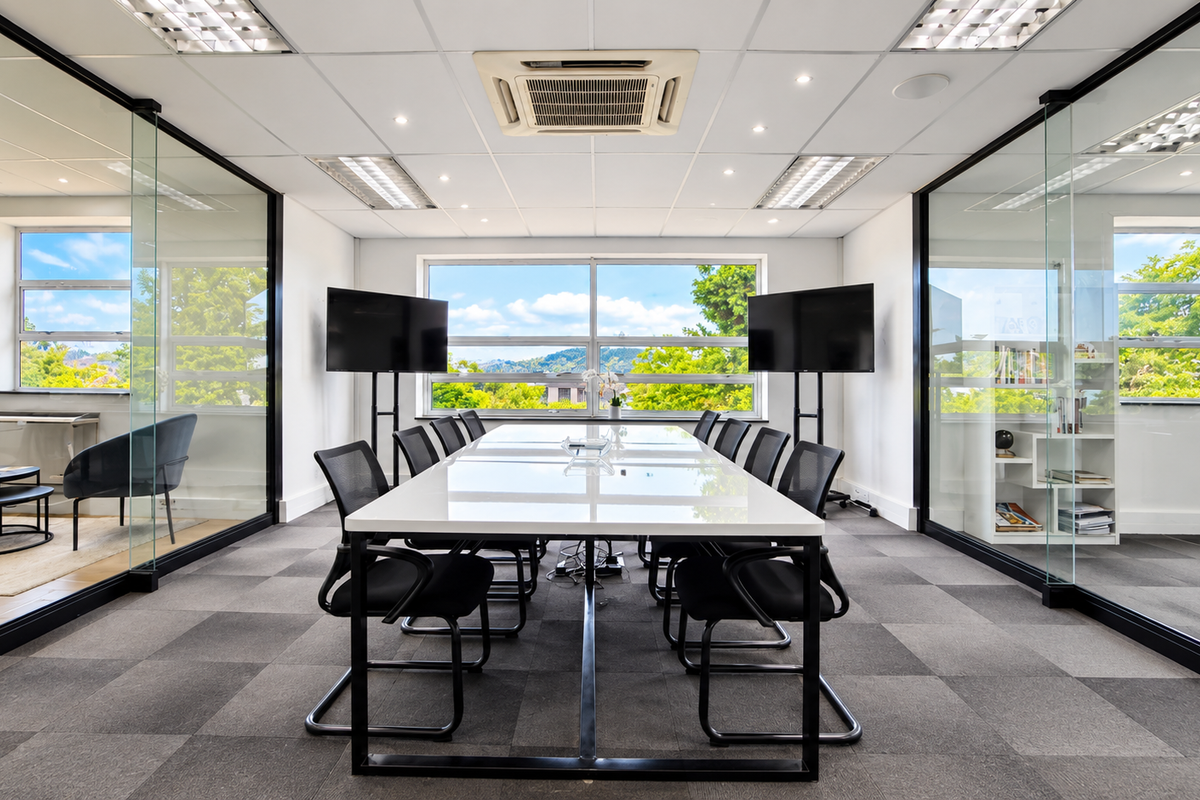 A modern, professional boardroom featuring glass partition walls, a suspended ceiling with integrated air conditioning, and carpeted flooring with ample natural light.