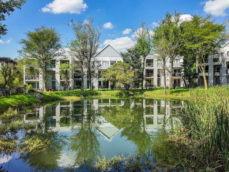 A scenic view of the apartment complex exterior featuring a tranquil pond in the foreground and well-maintained building facades with private balconies.