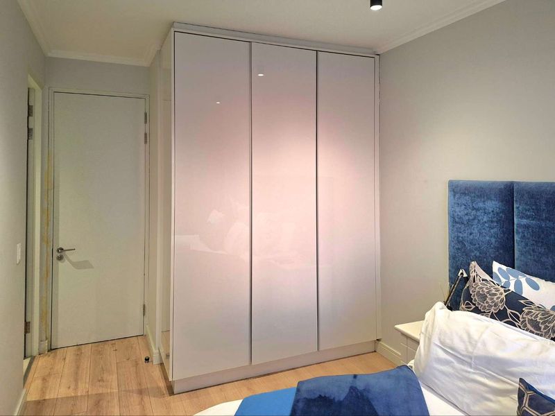 A bedroom featuring modern high-gloss built-in wardrobes, light laminate flooring, and a neutral color palette.