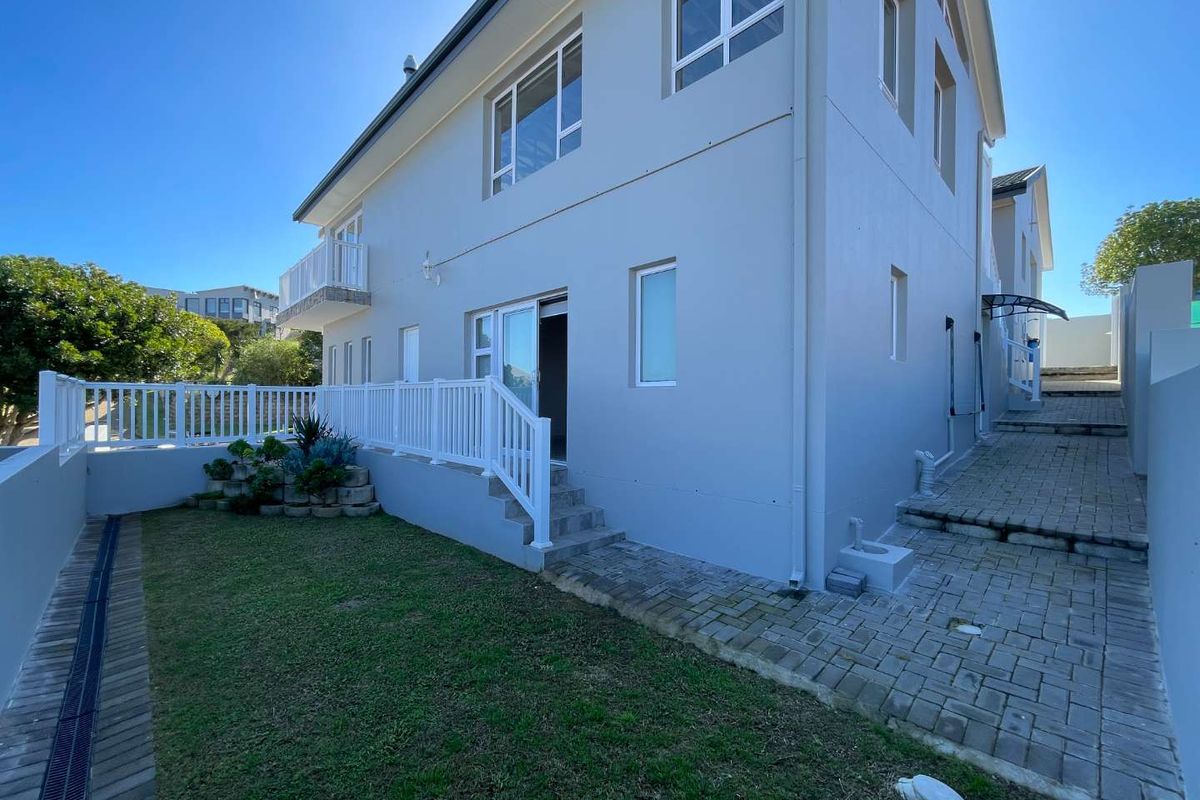 Rear exterior view of the property featuring a paved walkway, small lawn area, white balustrade, and access to the upper level balcony.