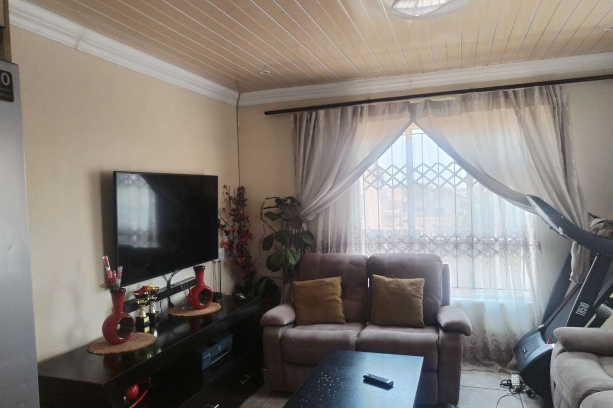 A comfortable living room featuring tiled flooring, a timber panelled ceiling, and a window fitted with a security gate.