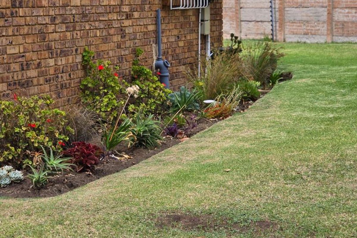 A side view of the townhouse exterior featuring a brick facade, security window bars, a solar panel installation, and a landscaped garden bed.