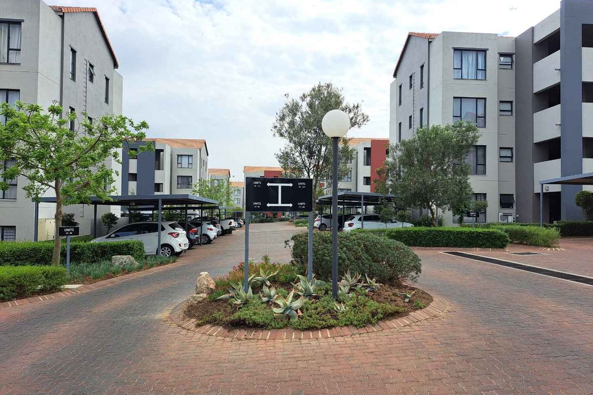 A well-maintained residential apartment complex featuring paved driveways, covered parking bays, and landscaped garden areas between multi-story building blocks.