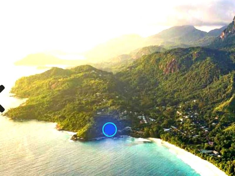 An aerial view of a coastal residential area featuring lush green mountains, a pristine white sand beach, and turquoise ocean waters.