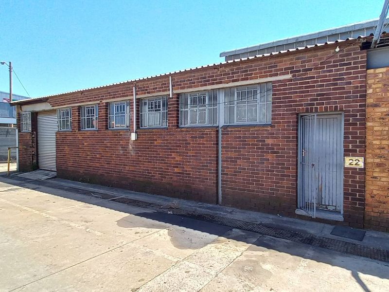 Exterior view of an industrial unit featuring brick facade, security window bars, and a steel roller shutter door with concrete driveway access.