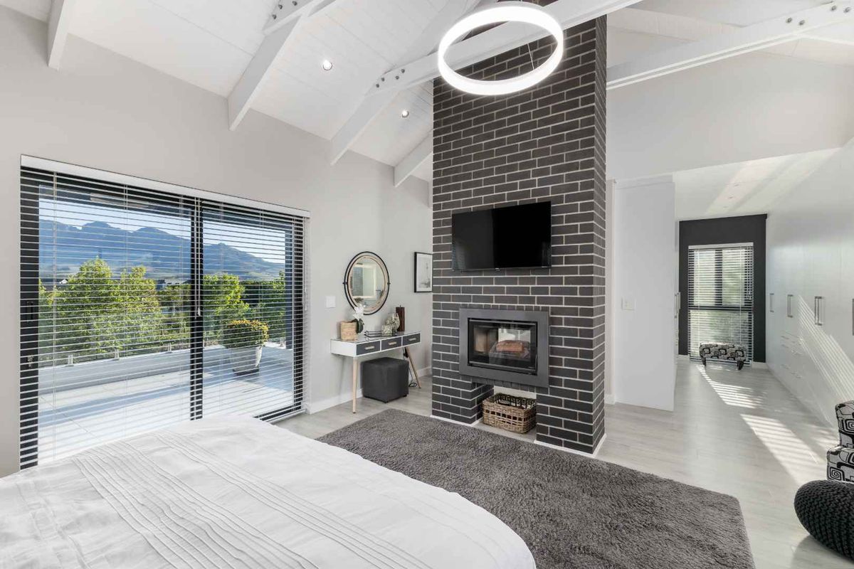 Spacious bedroom featuring a vaulted ceiling with exposed beams, a central brick fireplace, and large sliding glass doors opening to a balcony with mountain views.