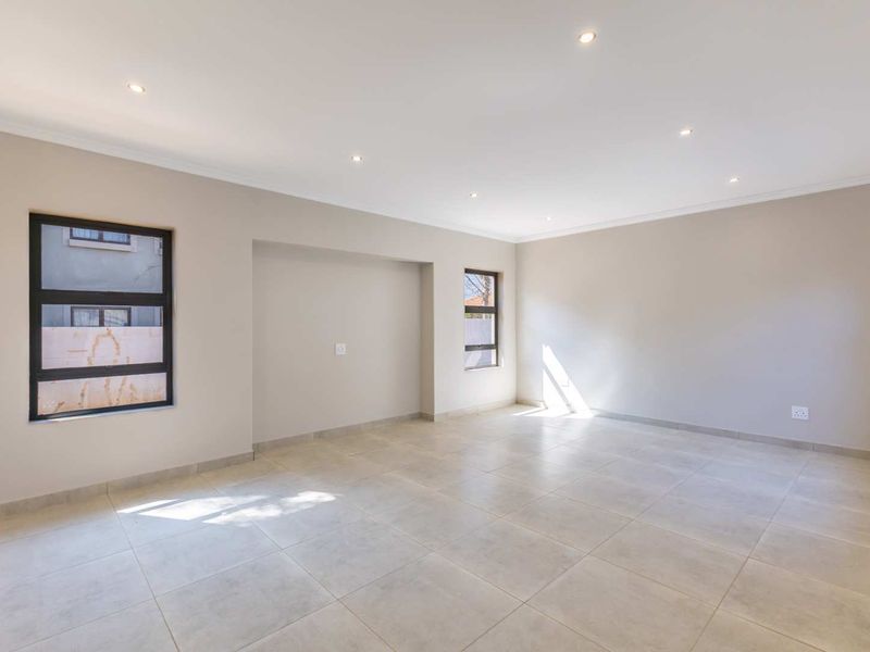 A bright, spacious, and empty living area featuring neutral wall tones, large aluminium-framed windows, and modern ceramic floor tiles with recessed downlighting.