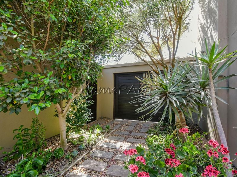 A well-maintained garden area featuring a stone paver pathway surrounded by mature trees, shrubs, and flowering plants against a boundary wall.