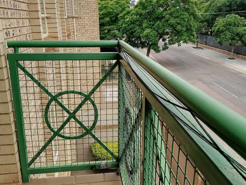A balcony featuring tiled flooring and a green metal balustrade, offering an elevated view of the street and surrounding trees.