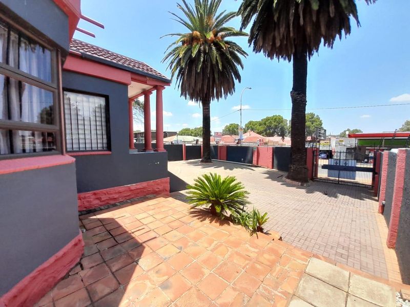 Front exterior view featuring a paved driveway, security gate, boundary wall, and a tiled patio area with established palm trees.