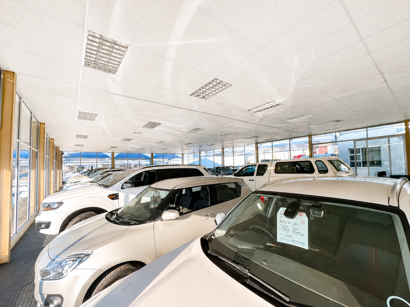 A spacious retail showroom featuring a suspended ceiling with recessed lighting, large glass windows, and durable rubberized flooring, currently utilized for vehicle display.