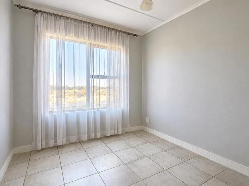 A bright, empty bedroom featuring neutral tiled flooring, light-colored walls, and a large window with sheer curtains.