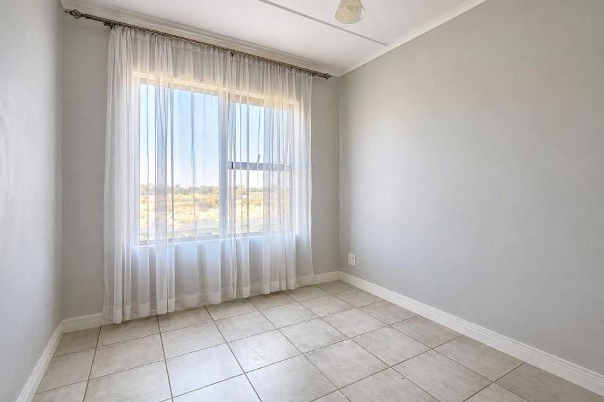A bright, empty bedroom featuring neutral tiled flooring, light-colored walls, and a large window with sheer curtains.