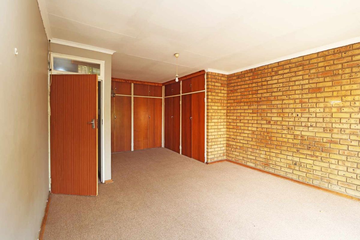An empty bedroom featuring an exposed brick feature wall, built-in wooden cupboards, and carpeted flooring, offering a blank canvas for renovation.