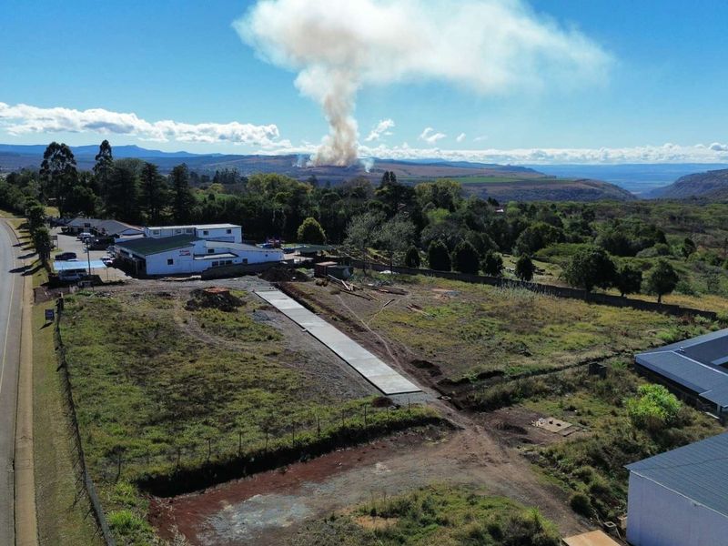Aerial view of a commercial vacant land plot featuring a concrete driveway, perimeter fencing, and adjacent commercial buildings with surrounding landscape.