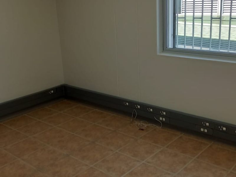 An empty office space featuring tiled flooring, integrated skirting trunking for power and data connectivity, and a window fitted with security bars.