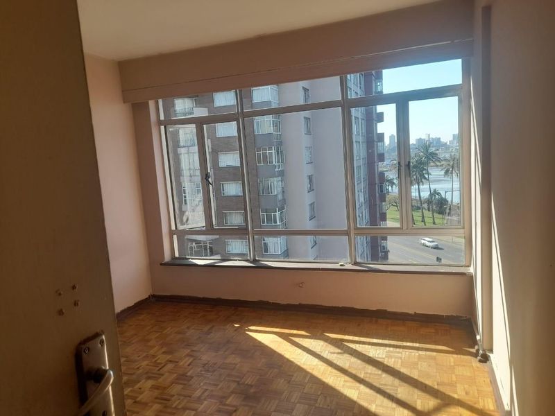 An empty bedroom featuring original parquet flooring and large windows offering urban and coastal views, ready for renovation or staging.