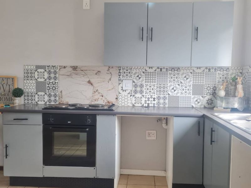 A functional kitchen featuring grey cabinetry, patterned tile backsplash, built-in oven, electric hob, and a stainless steel sink with tiled flooring.