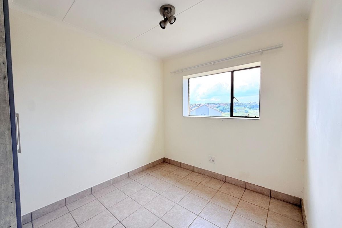 A clean, empty bedroom featuring neutral tiled flooring, a single window with natural light, and a ceiling-mounted spotlight fixture.