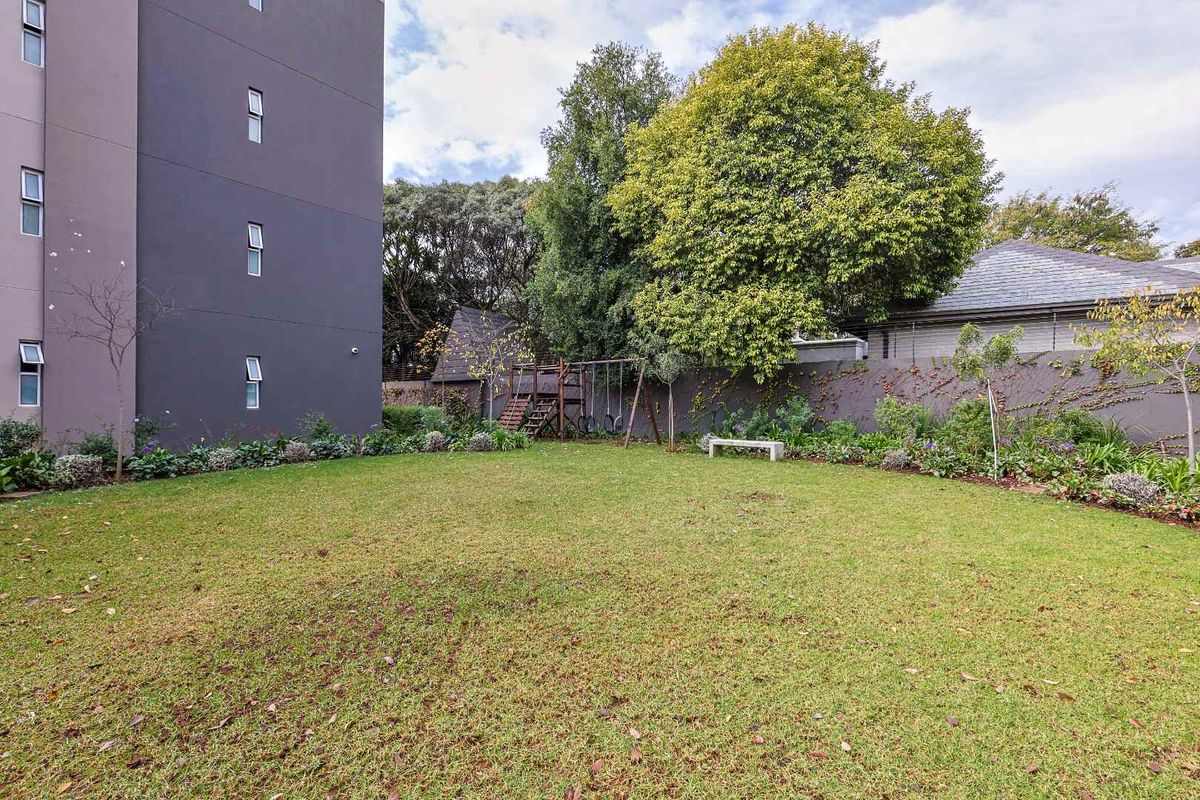 A well-maintained rear garden area featuring a spacious lawn, established trees, and perimeter boundary walling adjacent to the apartment building.