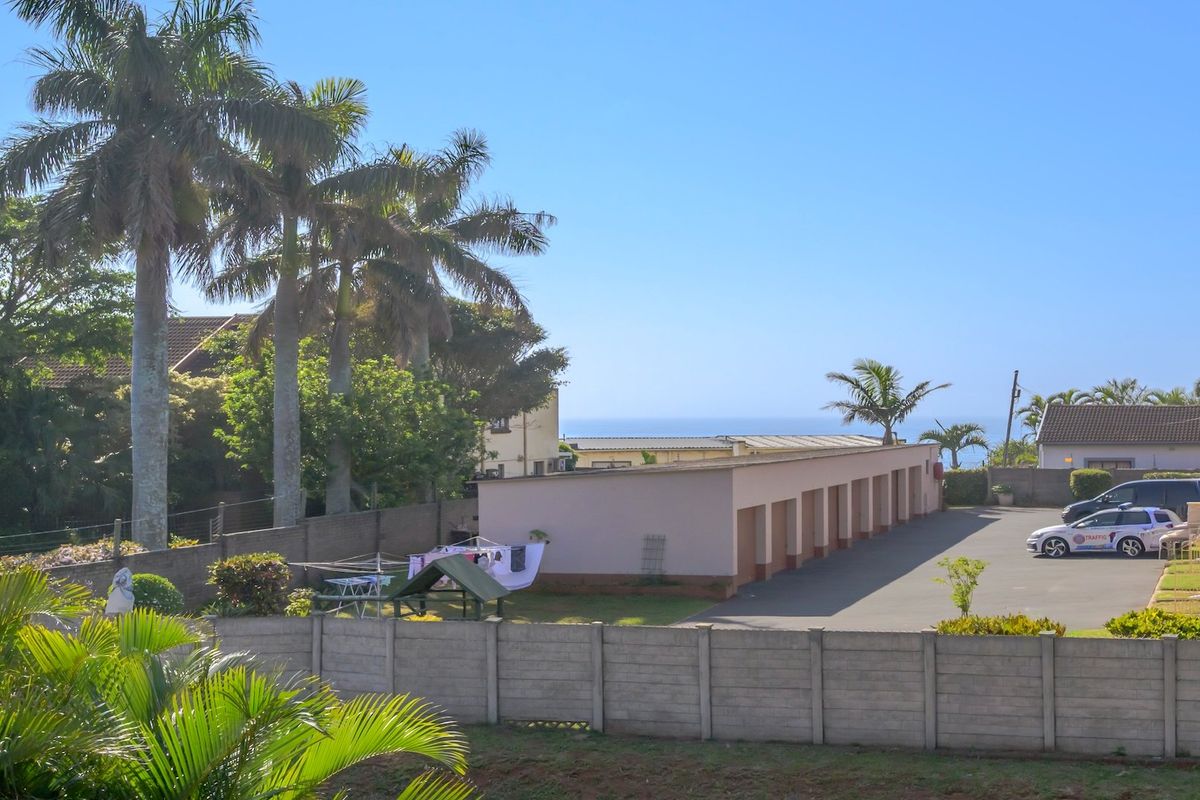 An elevated view of the apartment complex rear area, featuring a row of garages, paved driveway, and perimeter boundary wall with distant ocean views.