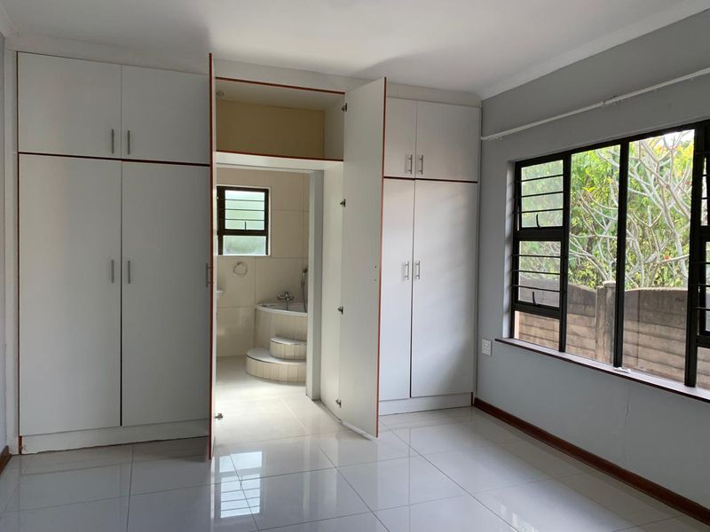 A bright, empty bedroom featuring tiled floors, extensive built-in cupboards, and direct access to an en-suite bathroom with a corner bath.