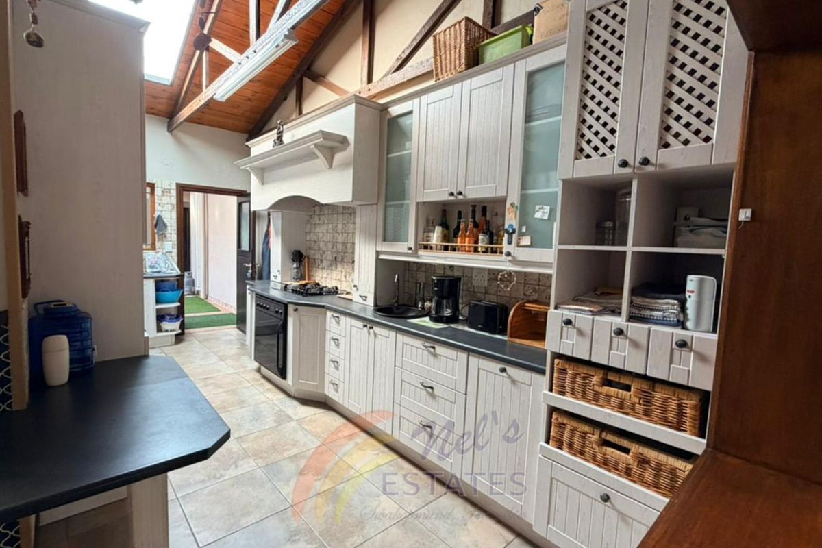 A rustic-style kitchen featuring timber cabinetry, a built-in oven and hob, exposed wooden roof trusses, and tiled flooring with access to an outdoor area.