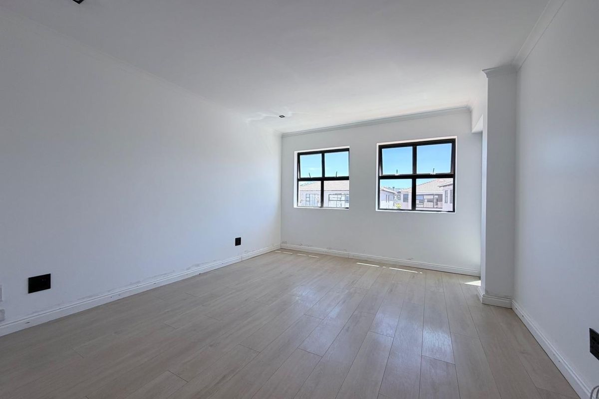 A bright, empty bedroom featuring light-toned laminate flooring, neutral walls, and black-framed windows providing natural light.