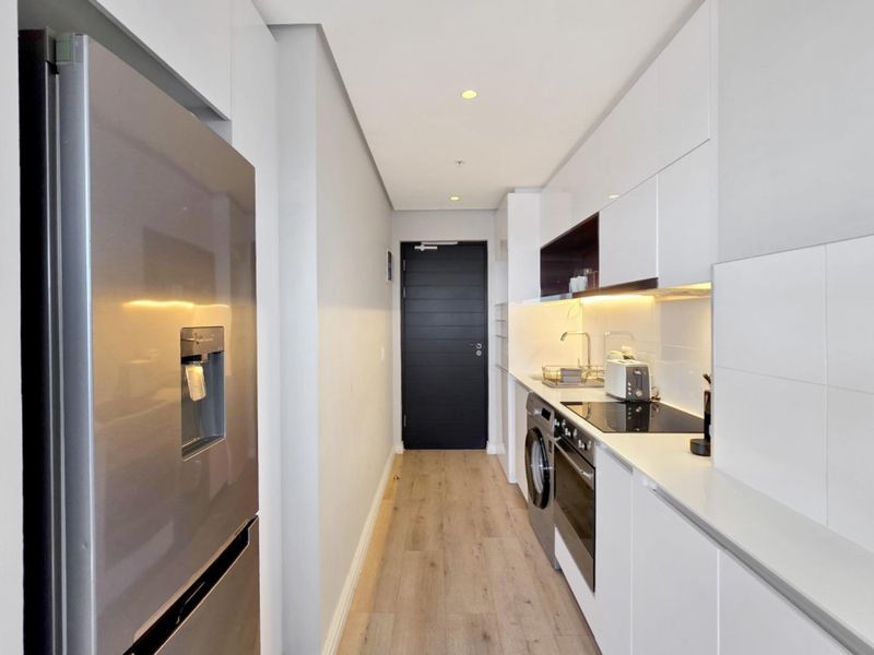 Modern galley-style kitchen featuring sleek white cabinetry, integrated appliances, electric hob, oven, and light wood-look flooring.
