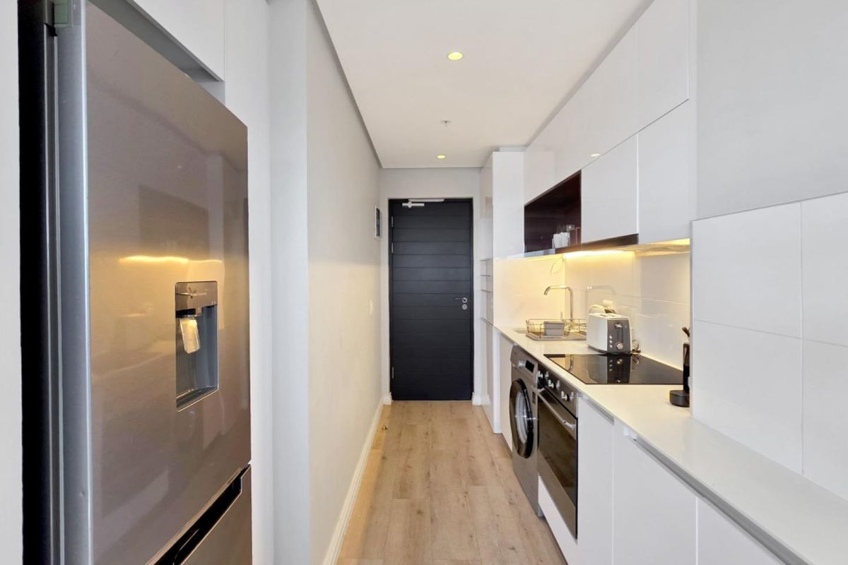 Modern galley-style kitchen featuring sleek white cabinetry, integrated appliances, electric hob, oven, and light wood-look flooring.