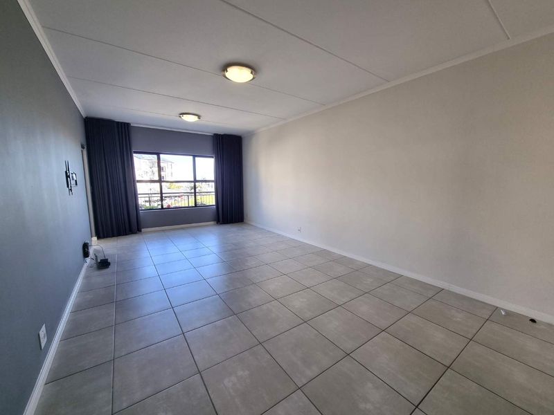 A spacious, empty living room featuring neutral tiled flooring, a large window with curtain tracks, and modern ceiling lighting.