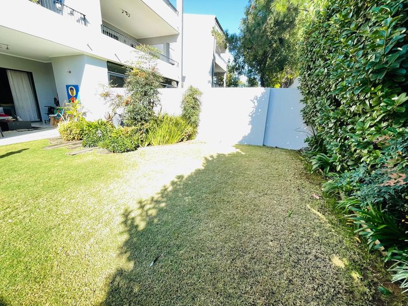 A private rear garden area featuring a well-maintained lawn, established perimeter hedging, and a boundary wall, adjacent to the apartment's patio.
