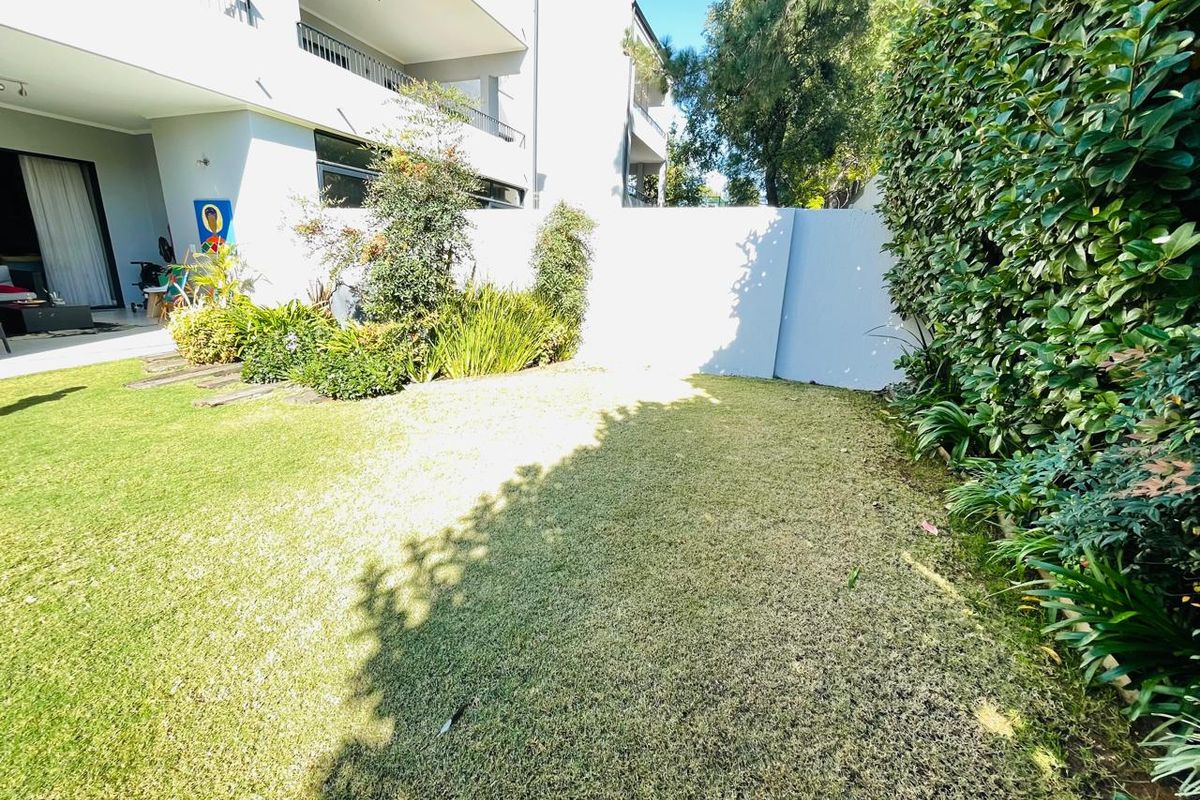 A private rear garden area featuring a well-maintained lawn, established perimeter hedging, and a boundary wall, adjacent to the apartment's patio.