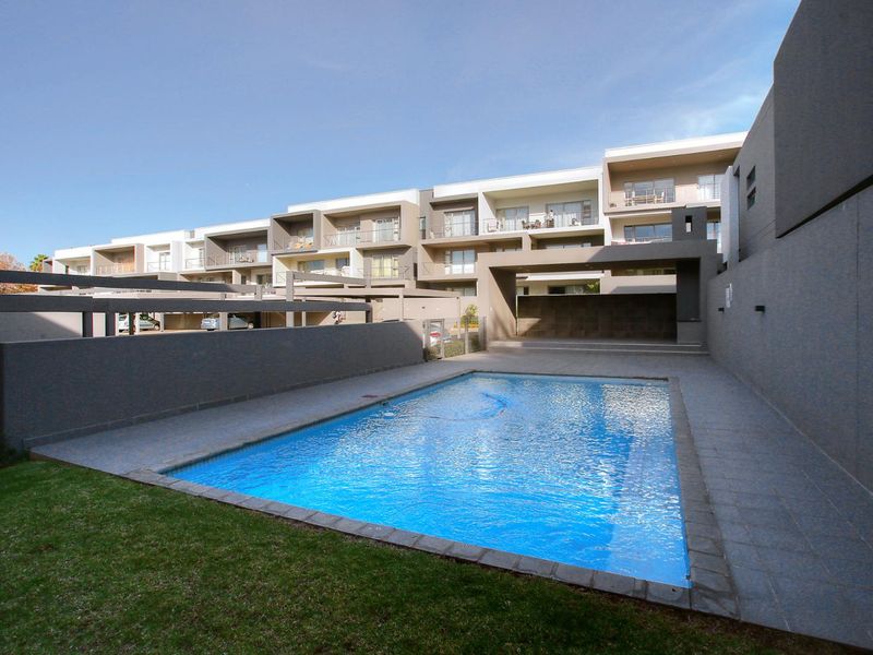 A well-maintained communal swimming pool area featuring a paved deck, covered patio space, and landscaped lawn within a modern apartment complex.