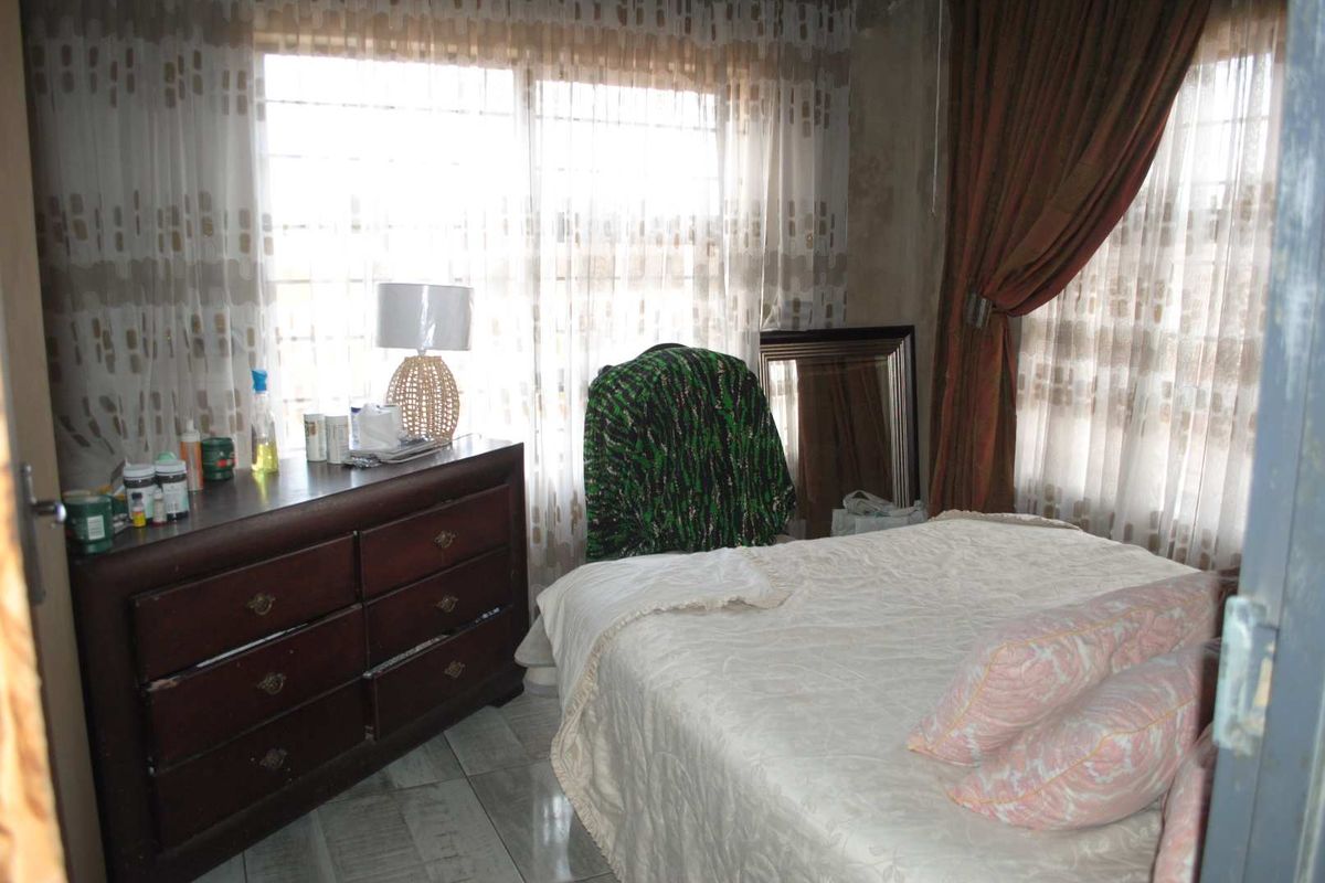 A bedroom featuring tiled flooring and large windows with curtains, showing signs of wear on the wall finishes.