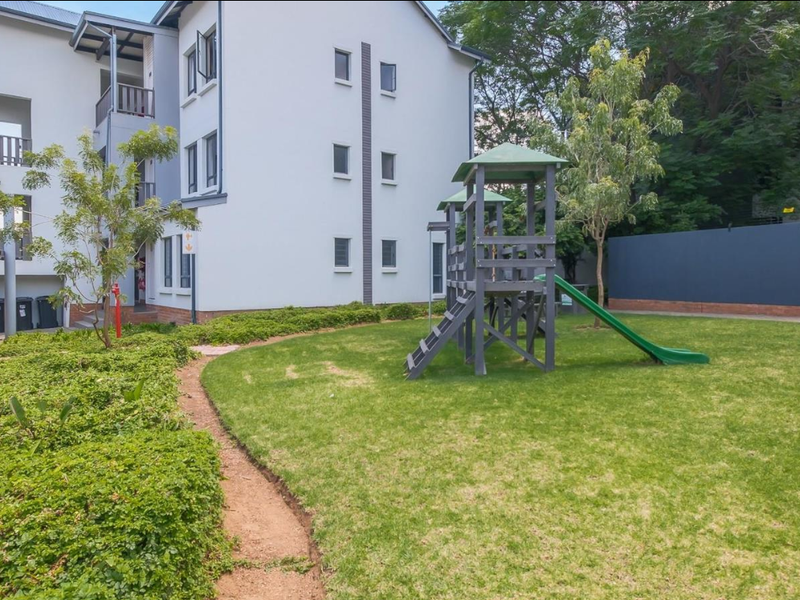 A well-maintained communal garden area featuring a children's play structure, manicured lawn, and paved pathways adjacent to the apartment building.