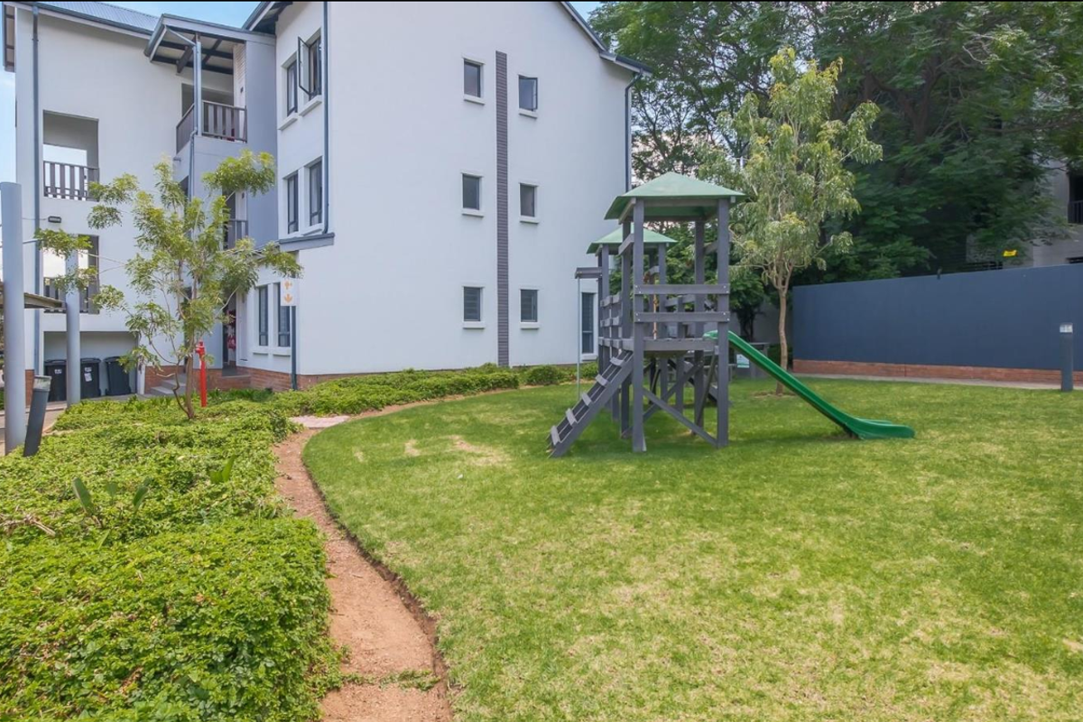 A well-maintained communal garden area featuring a children's play structure, manicured lawn, and paved pathways adjacent to the apartment building.
