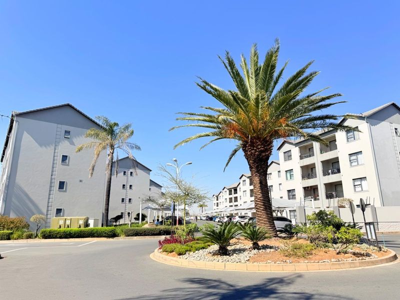 A view of the apartment complex entrance featuring a landscaped traffic circle, paved parking areas, and multi-story residential buildings under a clear sky.