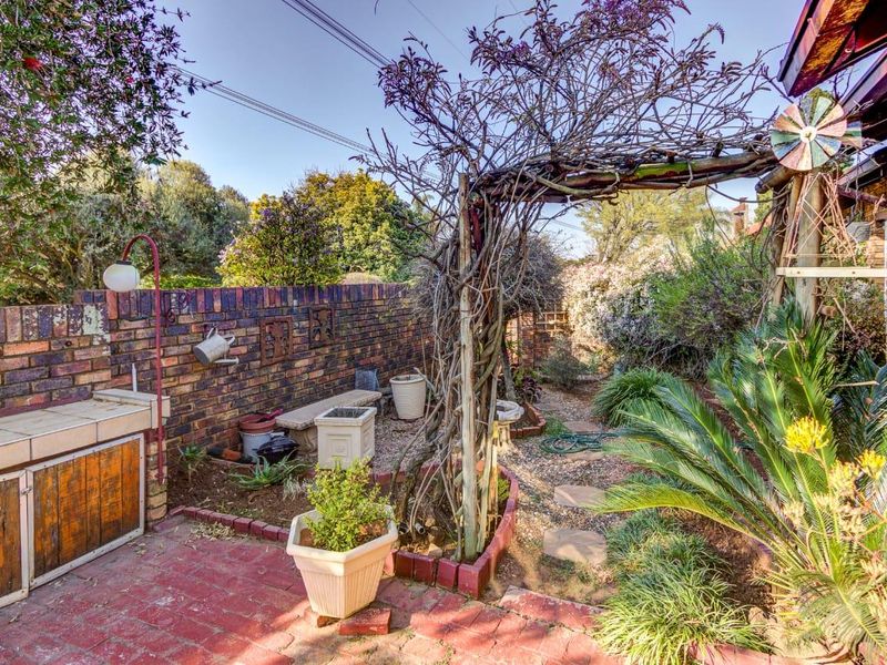A private rear garden area featuring a brick boundary wall, a built-in braai, a wooden pergola, and a stone pathway through landscaped beds.