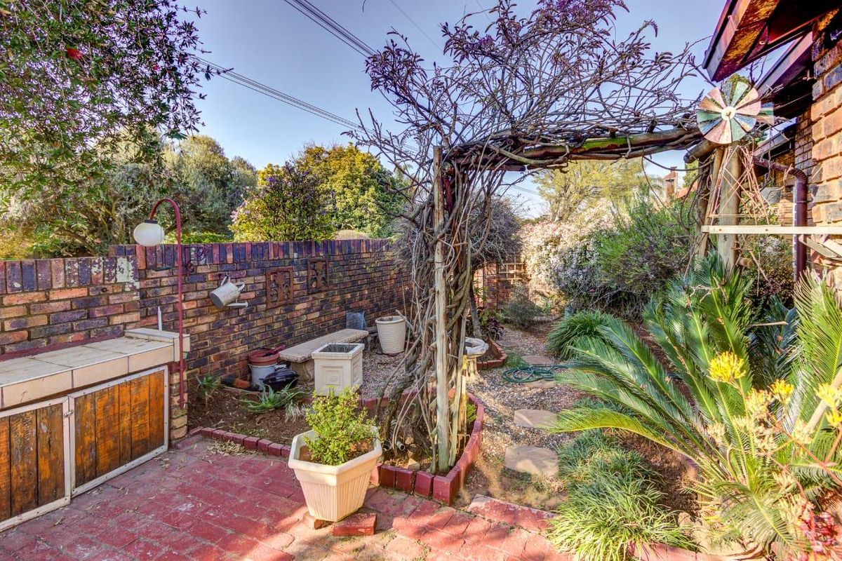 A private rear garden area featuring a brick boundary wall, a built-in braai, a wooden pergola, and a stone pathway through landscaped beds.